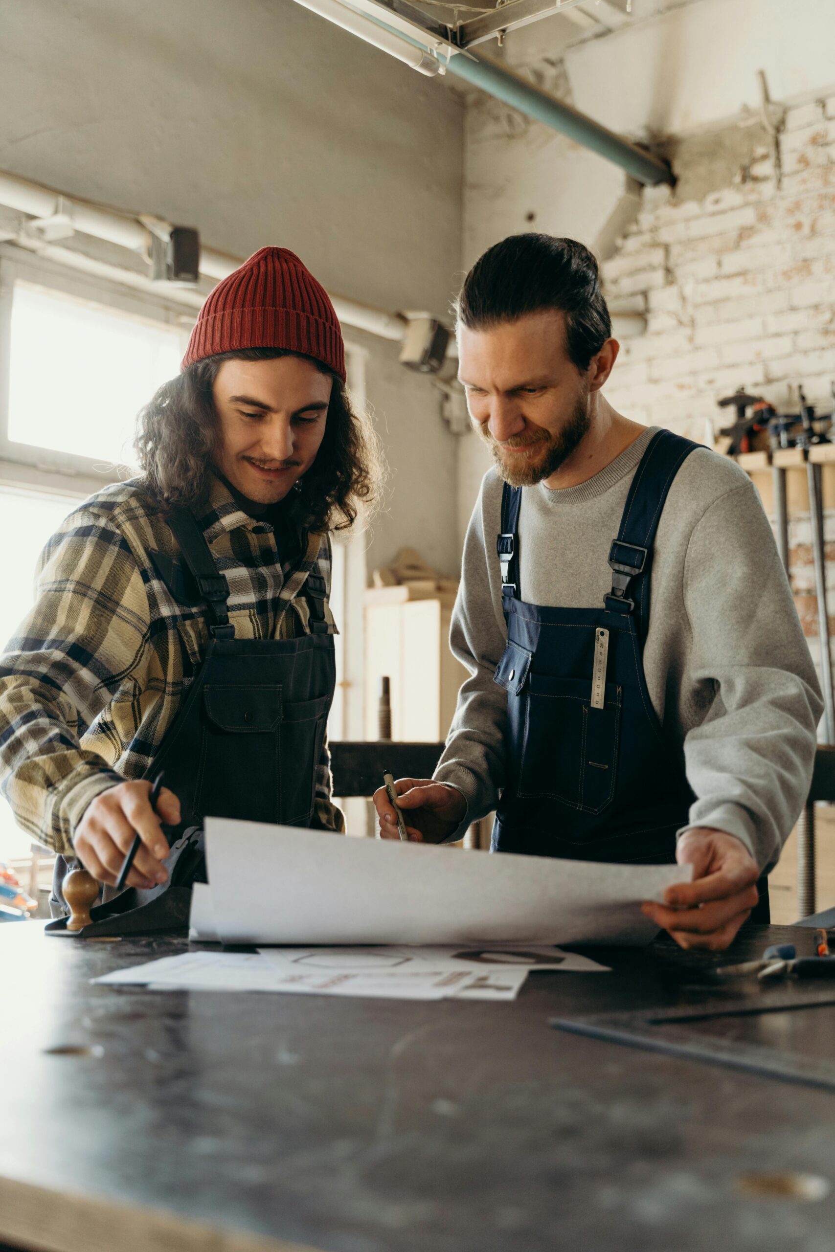 Two carpenters in overalls plan a project in an industrial workshop. Room is filled with tools and natural light.