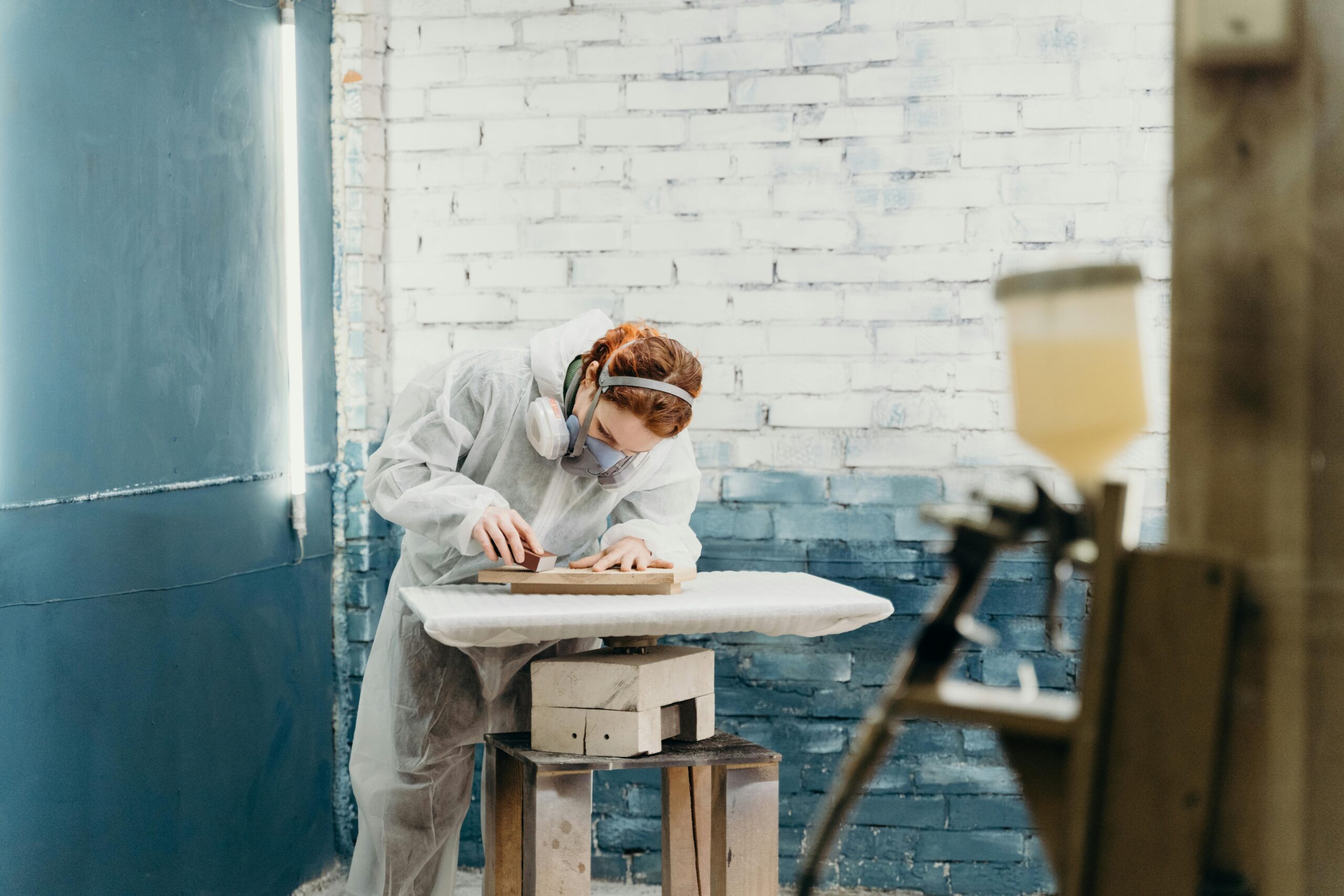 A craftsman in protective gear sands a wood piece in an industrial workshop.